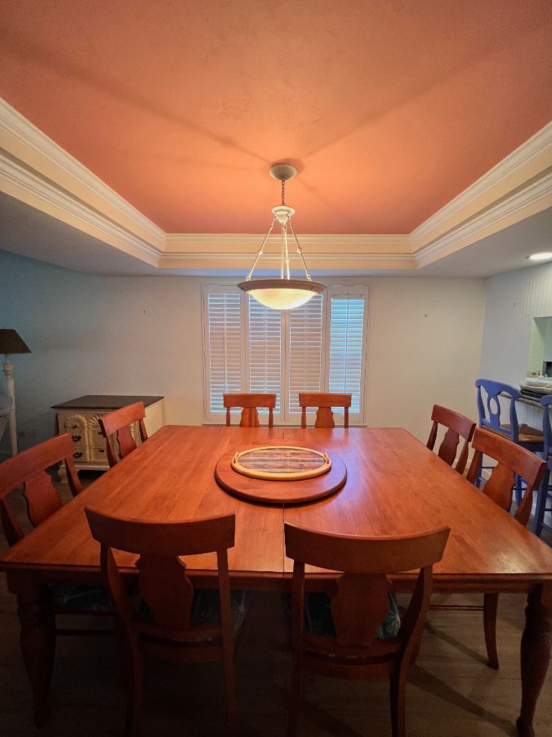 Dining room with wooden table, chairs, and pink ceiling. Window with shutters.