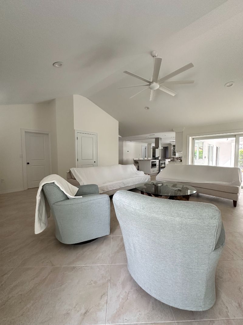 Spacious, light-filled living room with white walls, tiled floor, and soft blue armchairs. Two sofas are covered in white sheets.
