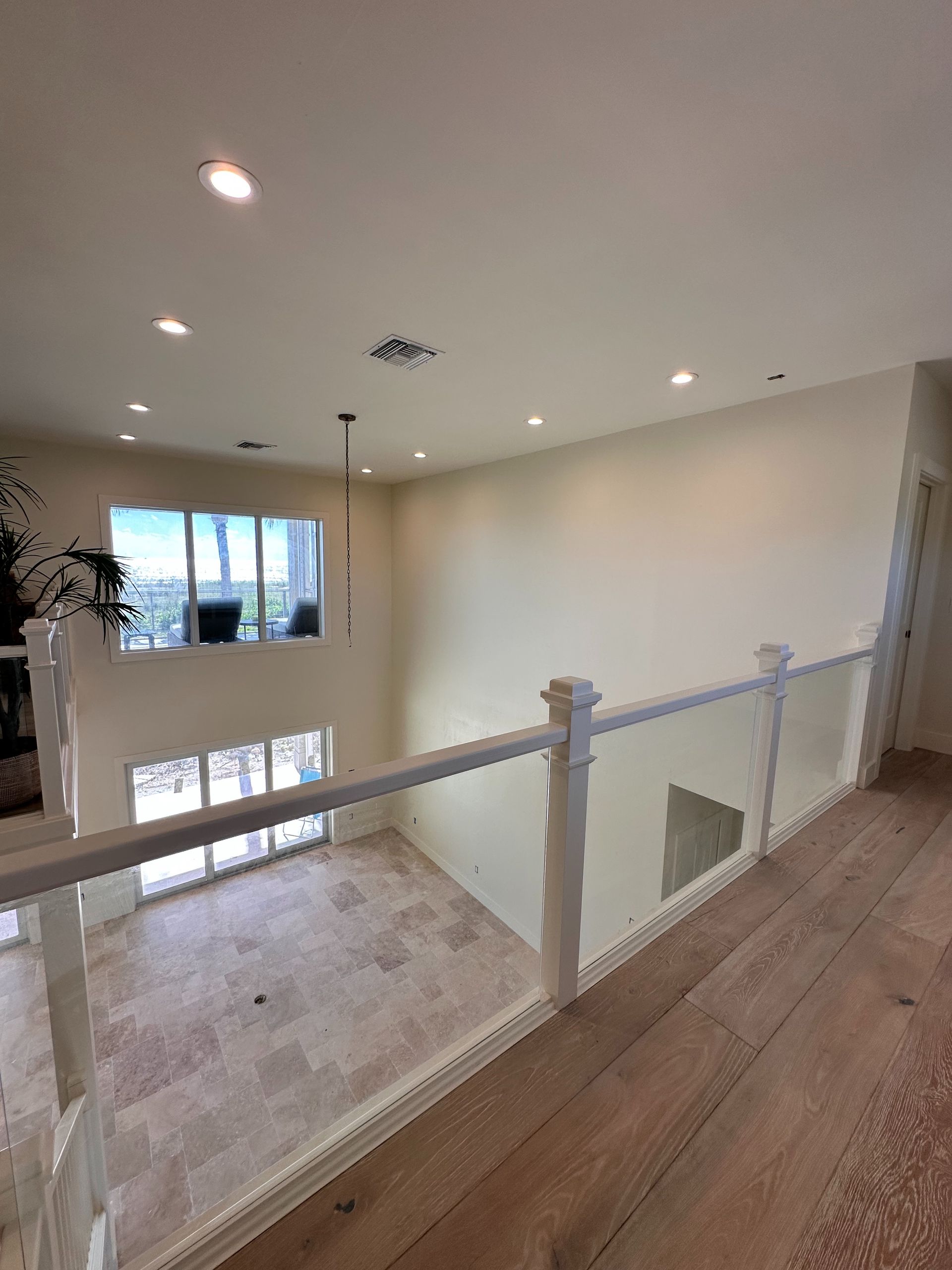 Interior view from a balcony overlooking a large living area with glass railings and tall windows.