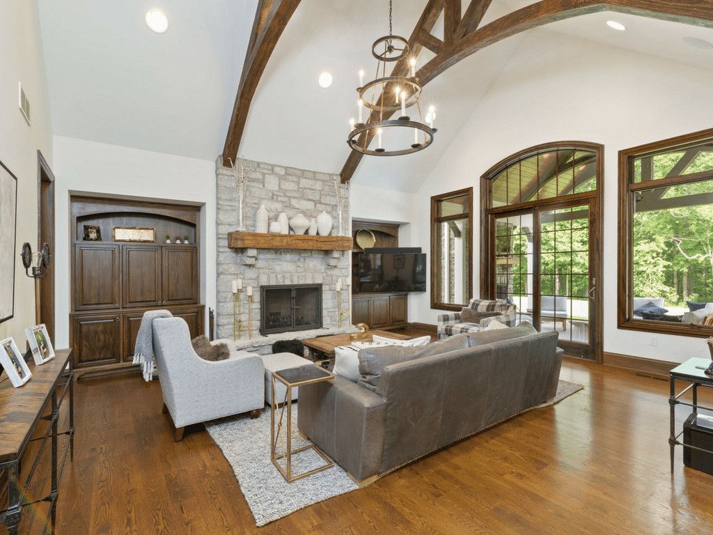 Living room with stone fireplace, exposed beams, and large windows overlooking greenery.