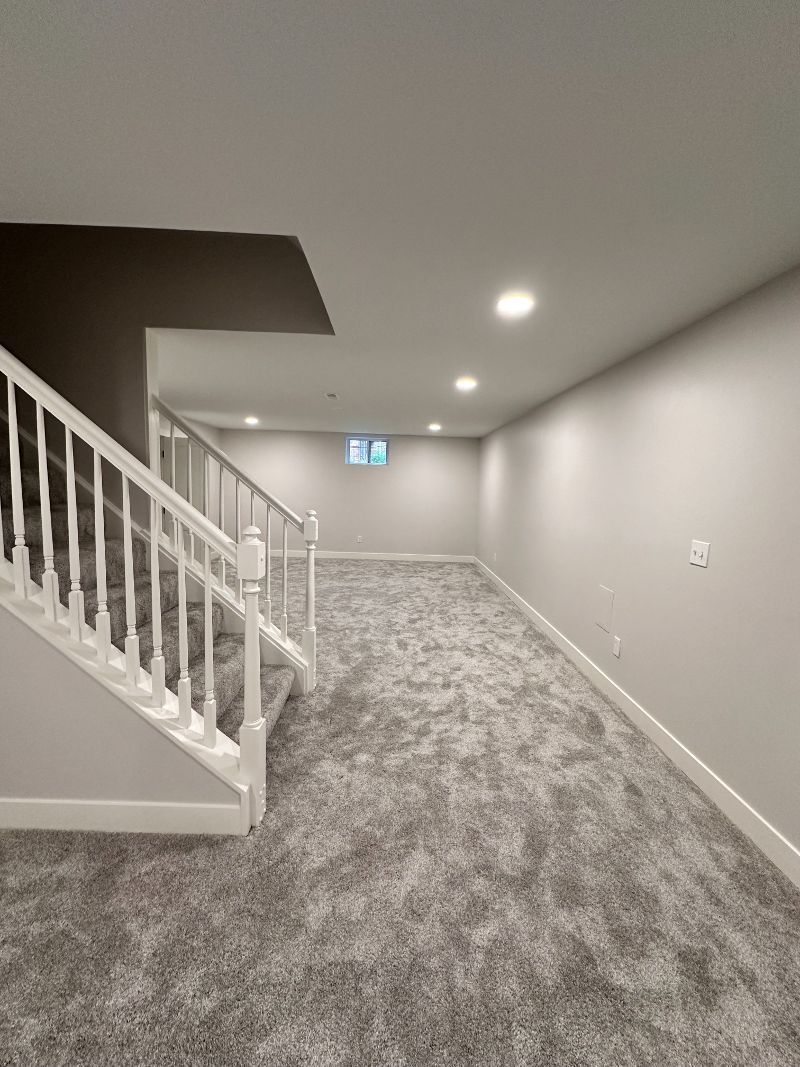 Basement with gray carpet, white stairs, and neutral walls. A small window is visible.