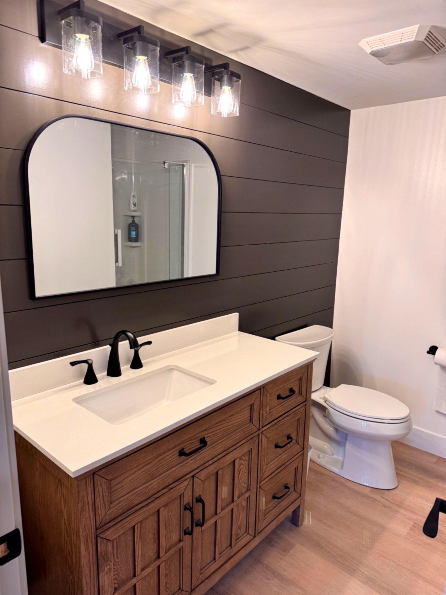 Bathroom with wood vanity, white countertop, black faucet, arched mirror, and a shiplap accent wall.