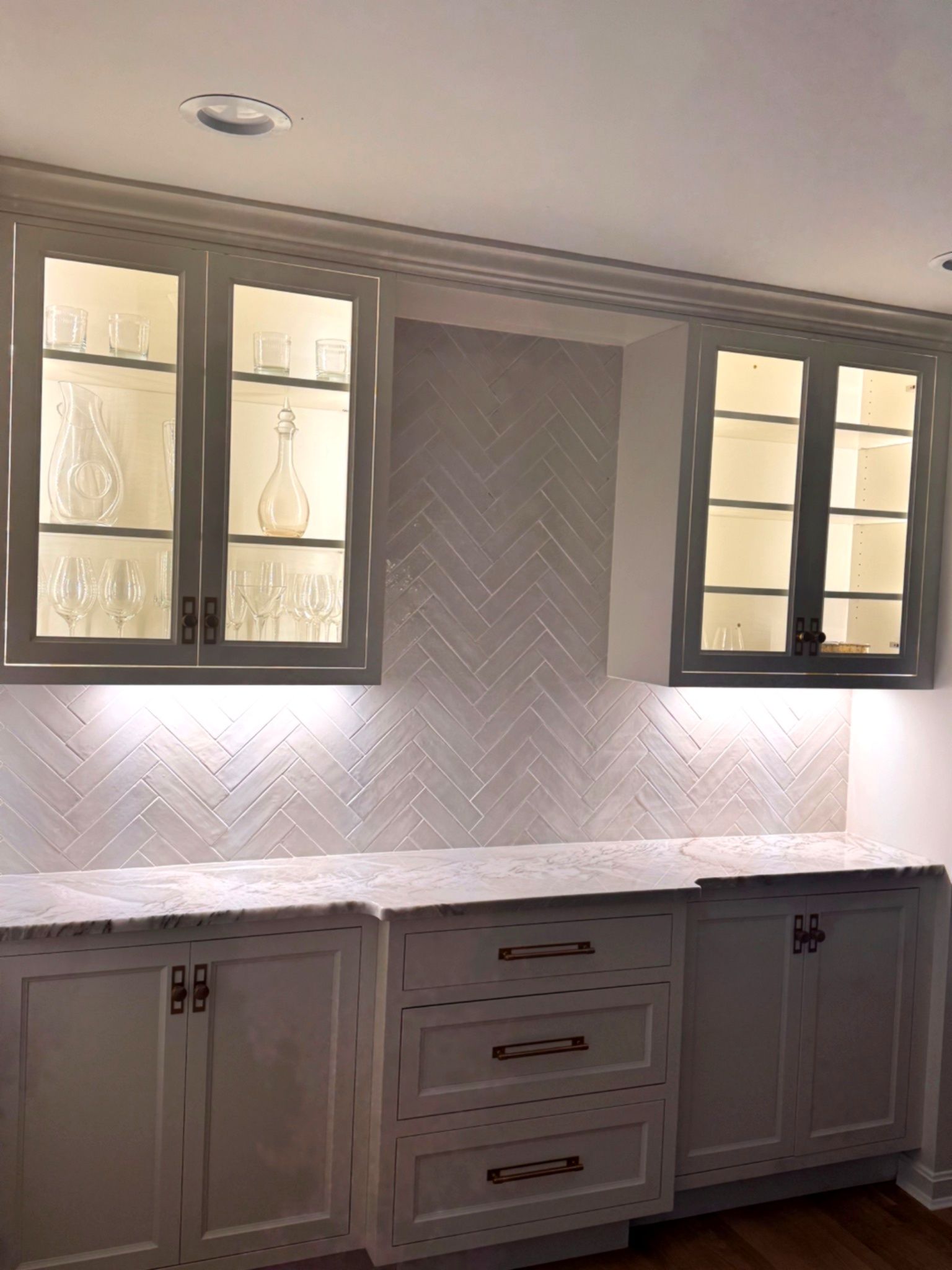 Kitchen cabinets with illuminated glass doors, white countertop, and herringbone backsplash.