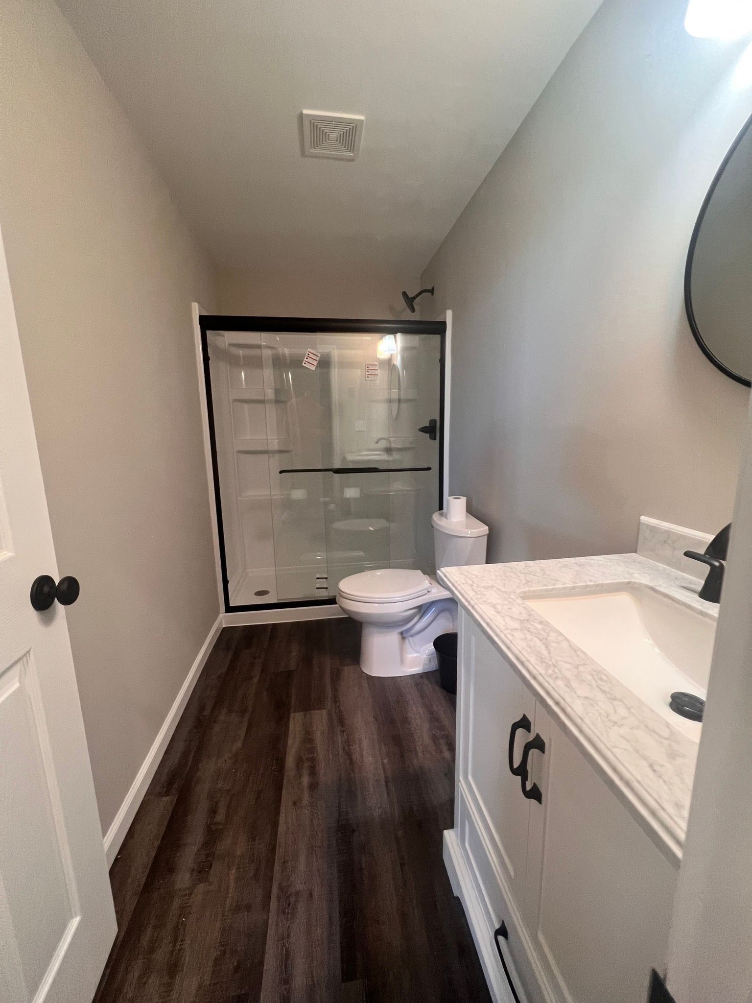 Narrow bathroom with dark wood-look flooring, white vanity, shower, toilet. Gray walls, black shower frame, and door hardware.