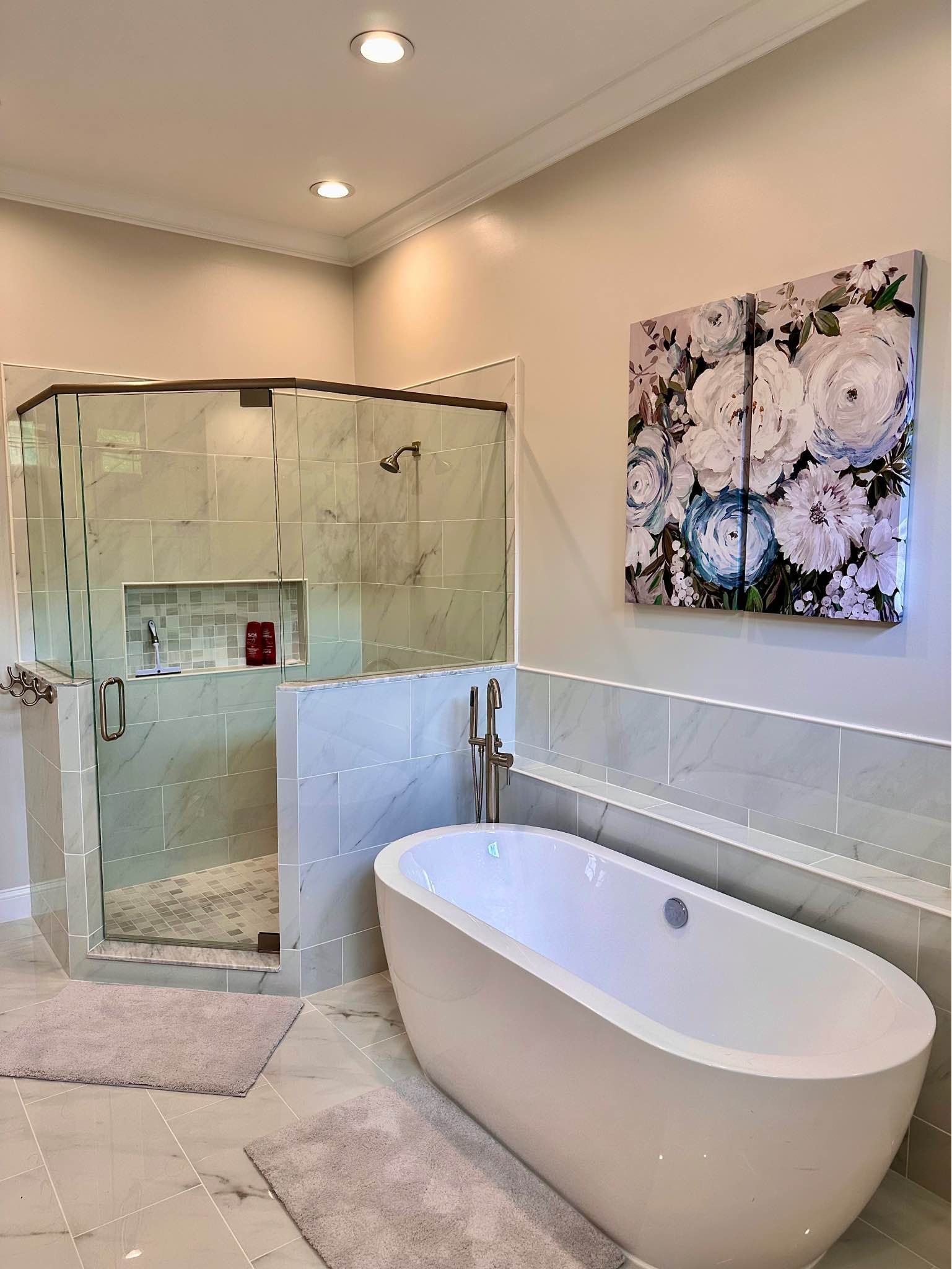 Bathroom with freestanding tub, glass shower, and floral artwork. Light gray walls and tile; white tub.