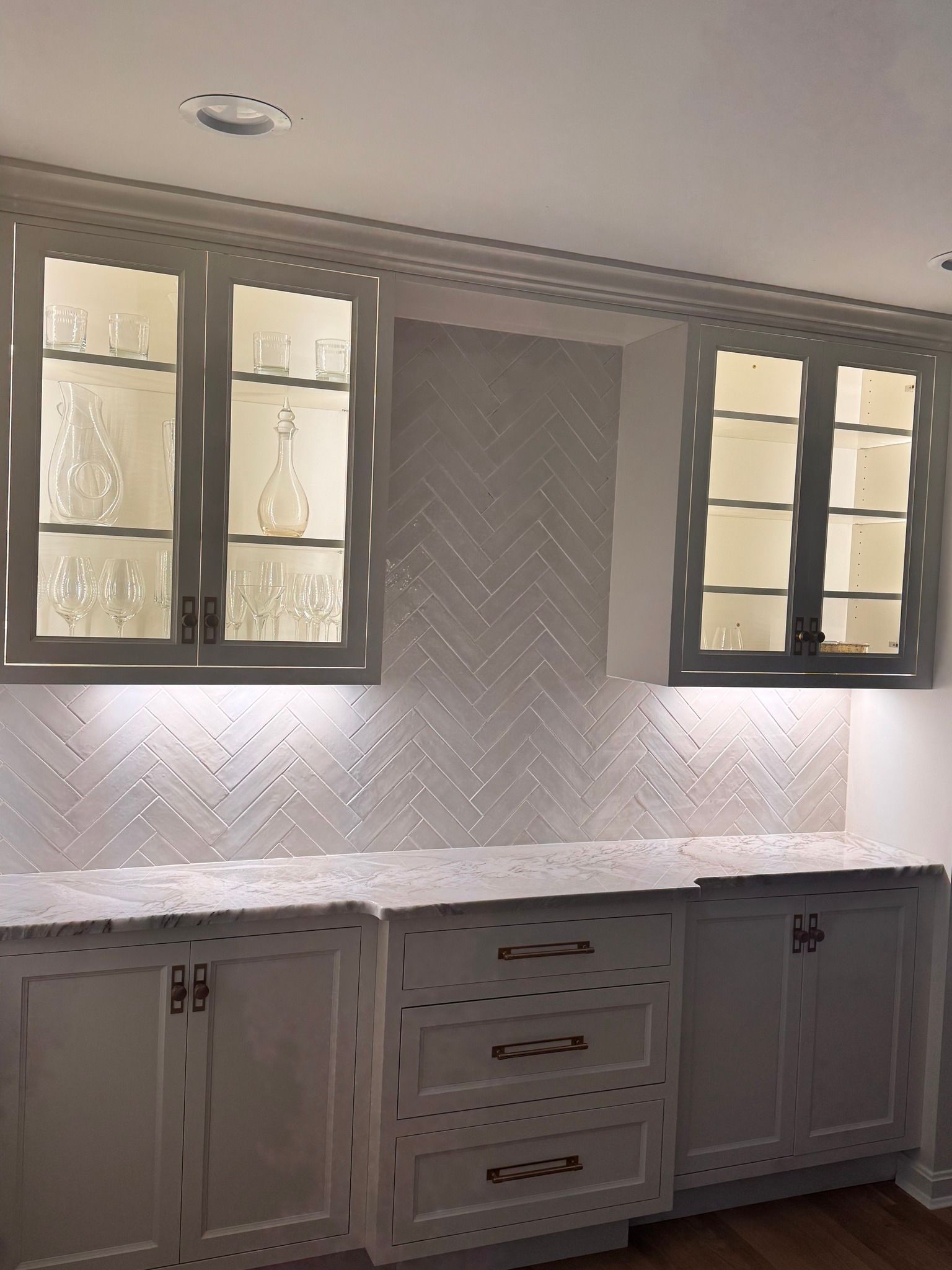 White built-in bar with glass-front cabinets displaying glassware, white herringbone backsplash, and white cabinets.
