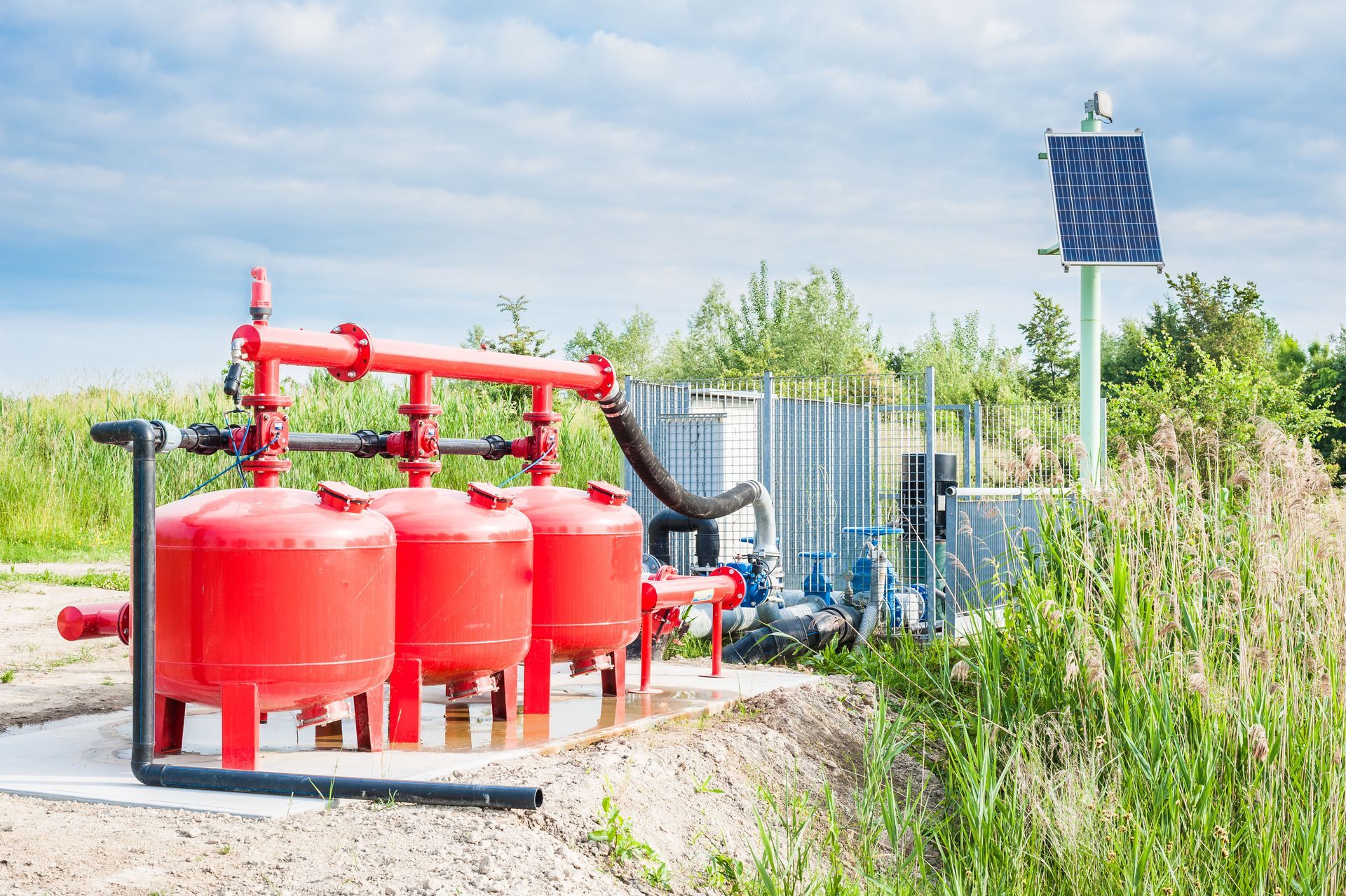 Red industrial tanks and pipes beside a solar-powered water treatment unit in a grassy field