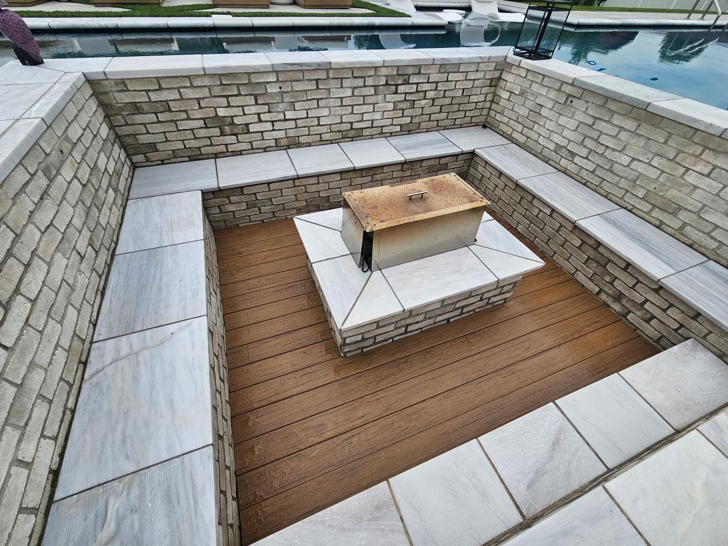 A sunken fire pit area with light stone seating, wooden decking, and a metal fire pit cover, adjacent to a swimming pool.