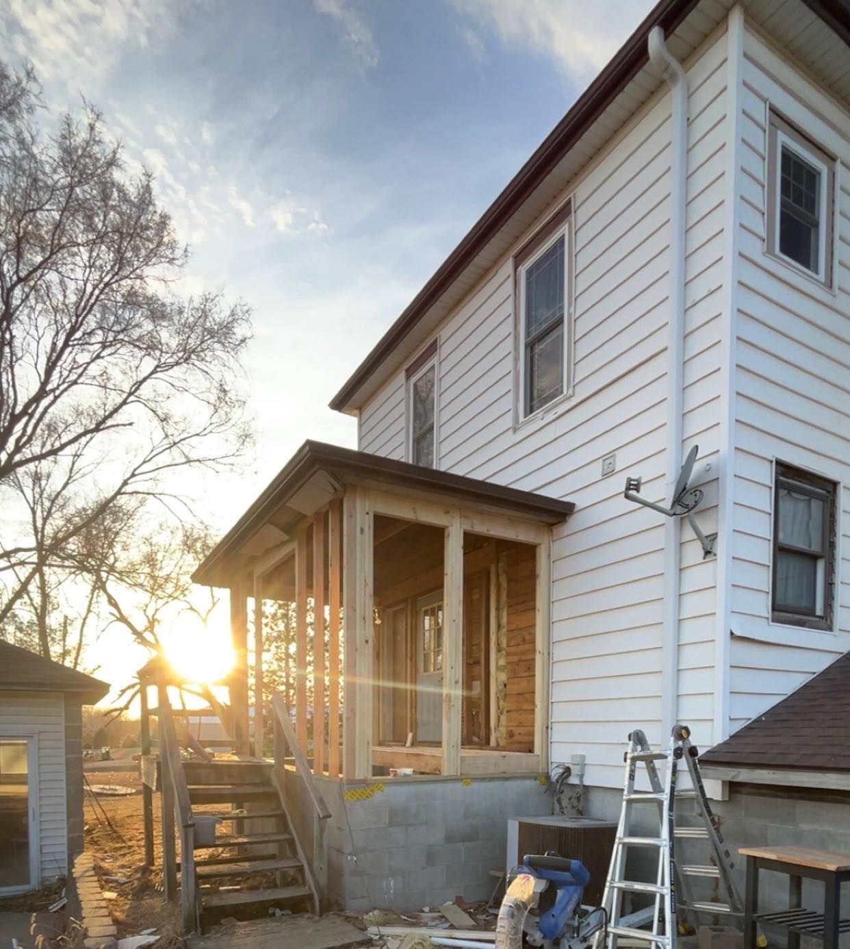 A wooden porch frame under construction against the white-sided wall of a house at sunset, with a ladder leaning nearby.