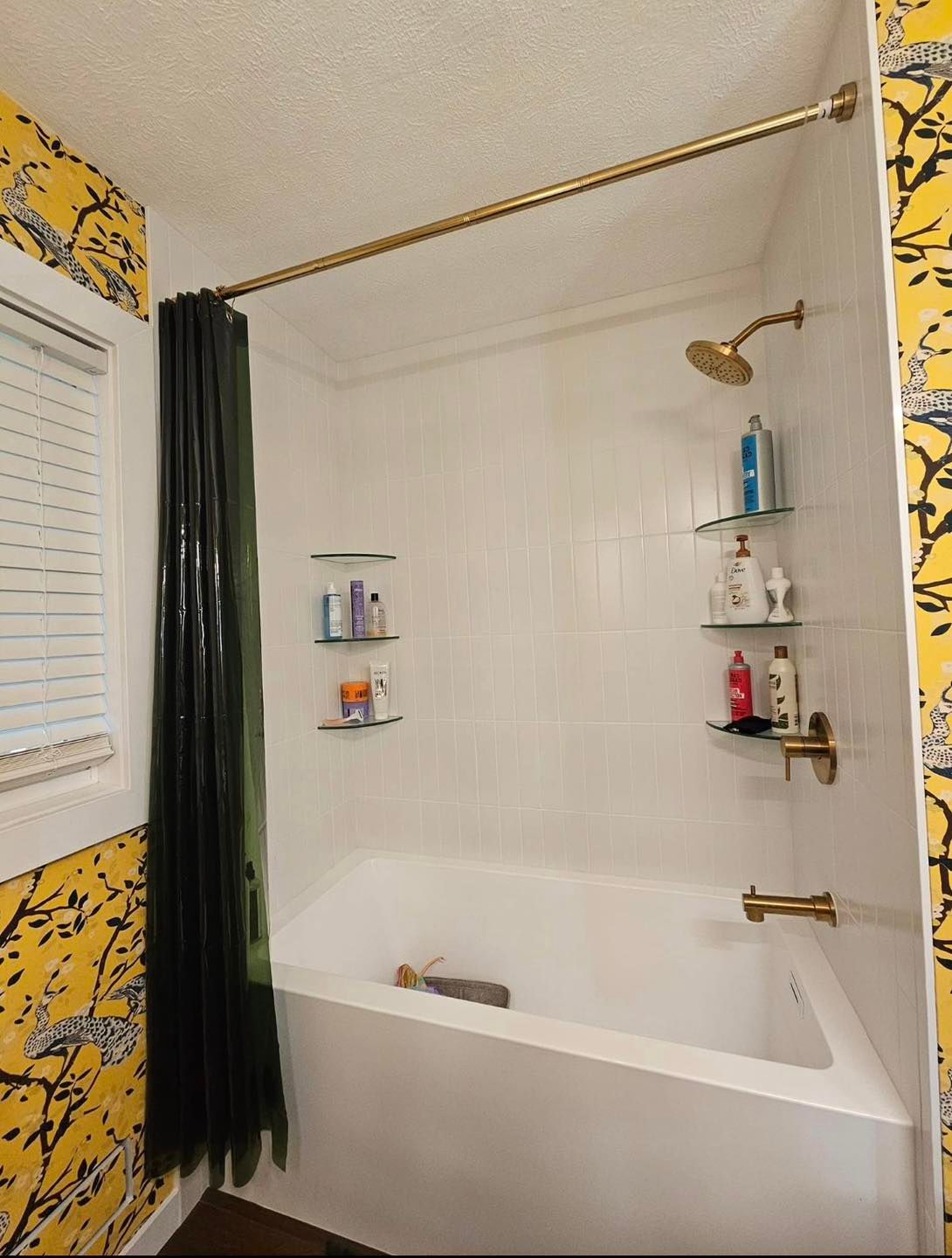 A bathtub with white walls, gold fixtures, two sets of glass corner shelves, and a green curtain against yellow wallpaper.