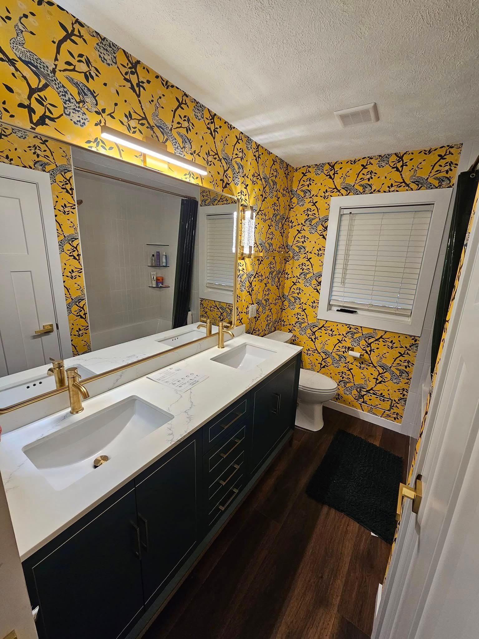 Bathroom with yellow patterned wallpaper, dark cabinets, a white double vanity, and wood-look flooring.