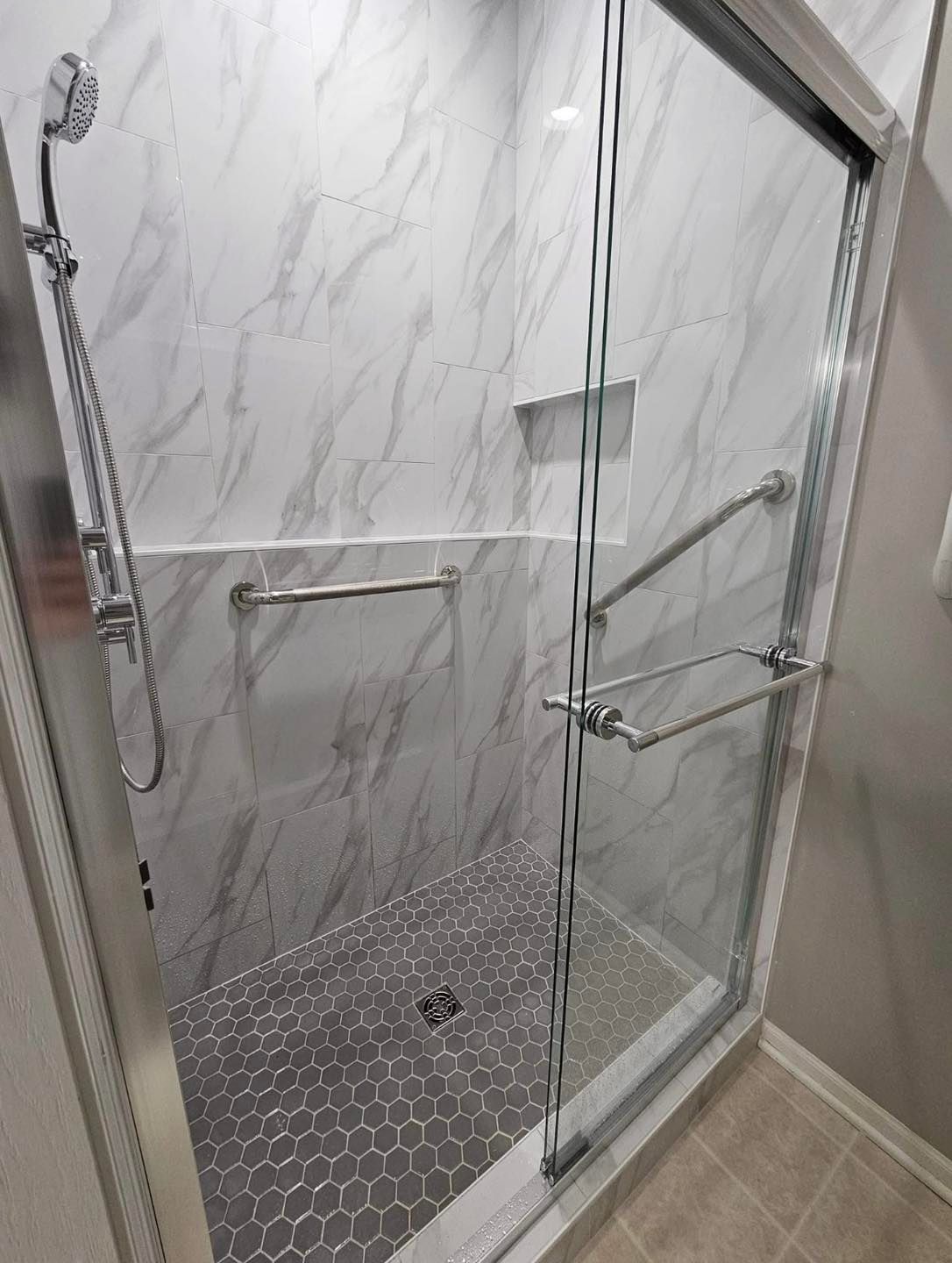 A walk-in shower with white marble-patterned walls, grey hexagon tile floors, a handheld showerhead, and safety grab bars.