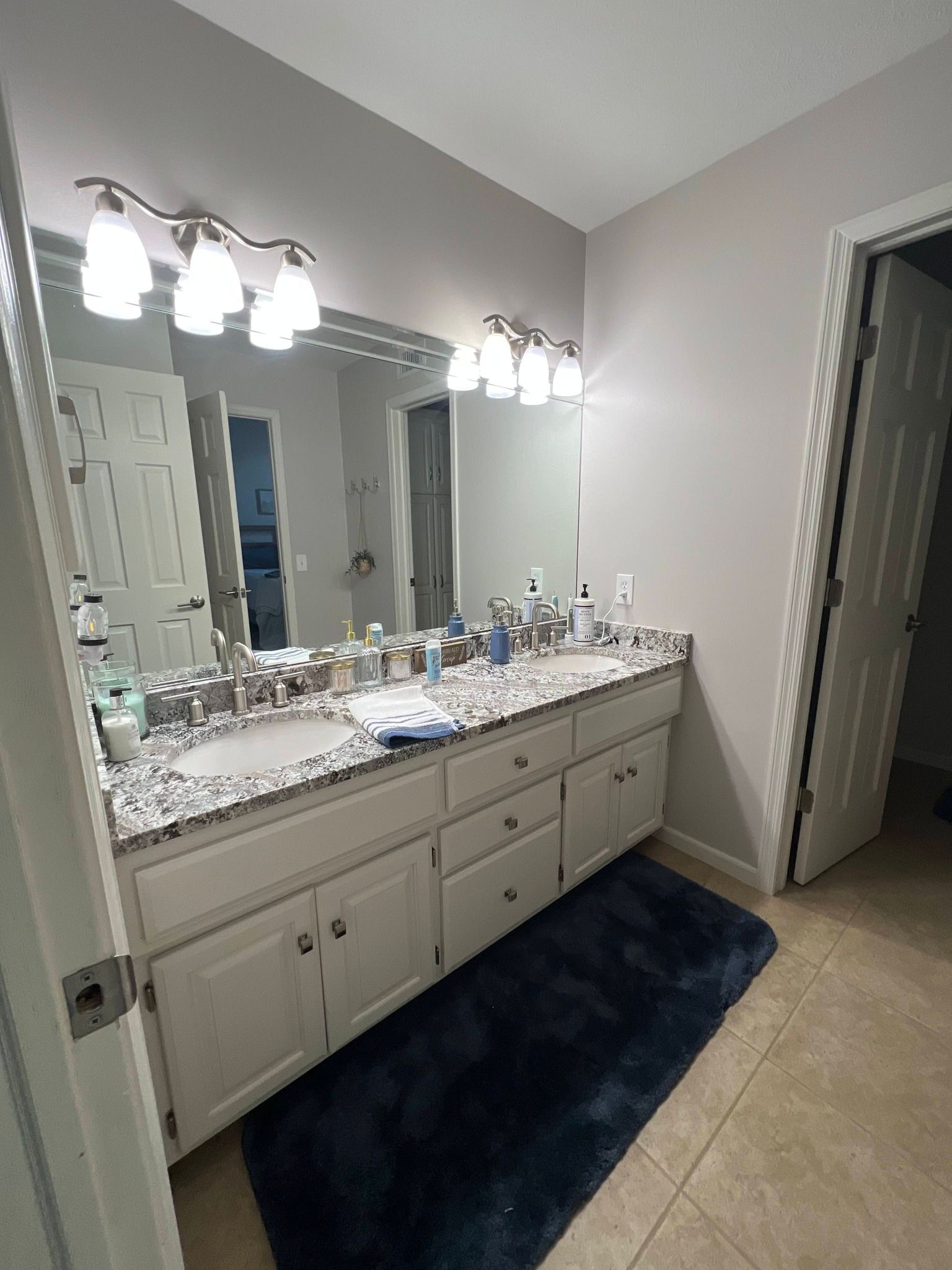 A bathroom with a double-sink vanity, marble-patterned countertop, white cabinets, two vanity lights, and a dark blue rug.