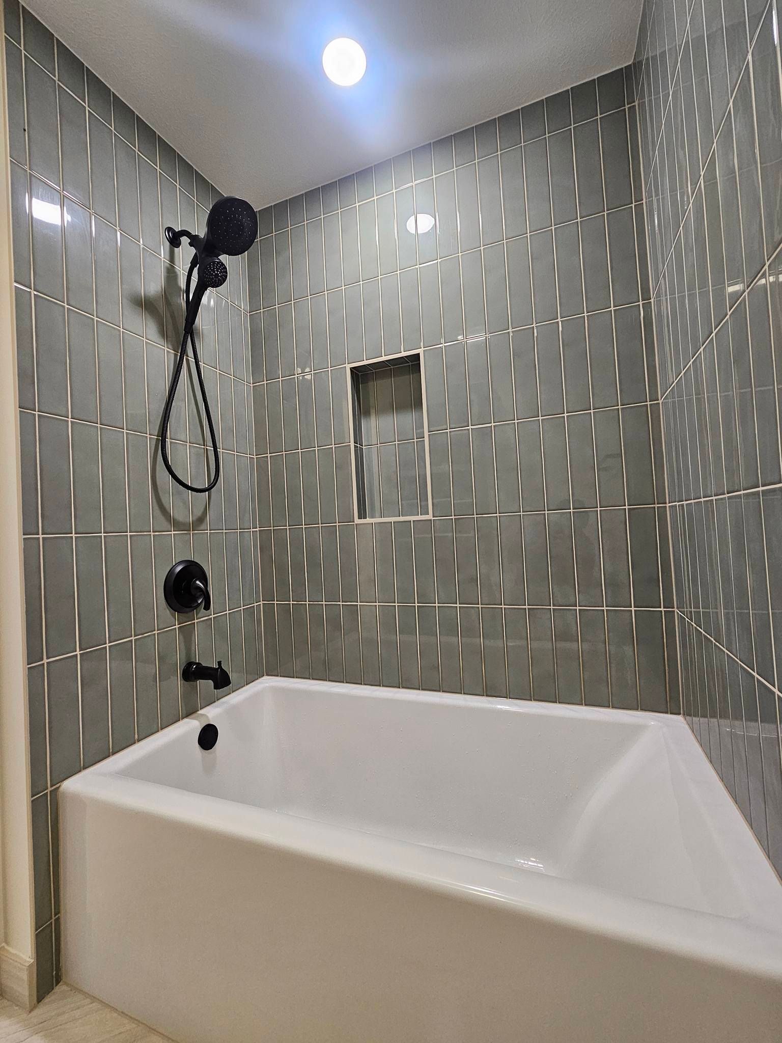A white bathtub with matte black fixtures set against walls with vertical, grey, rectangular tiles and a small niche.