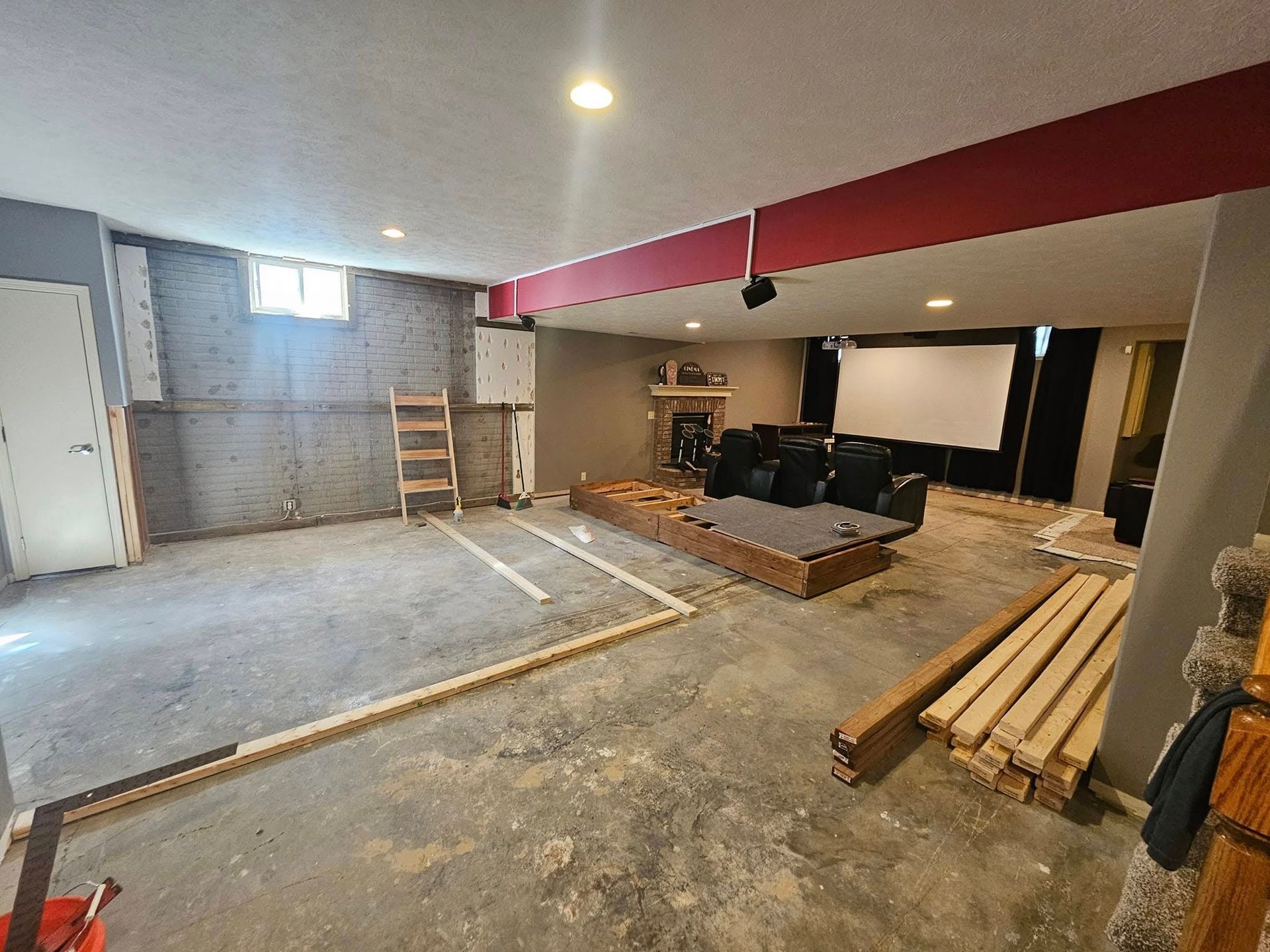 A basement undergoing renovation with a raised seating platform, a projector screen, and construction materials on the floor.