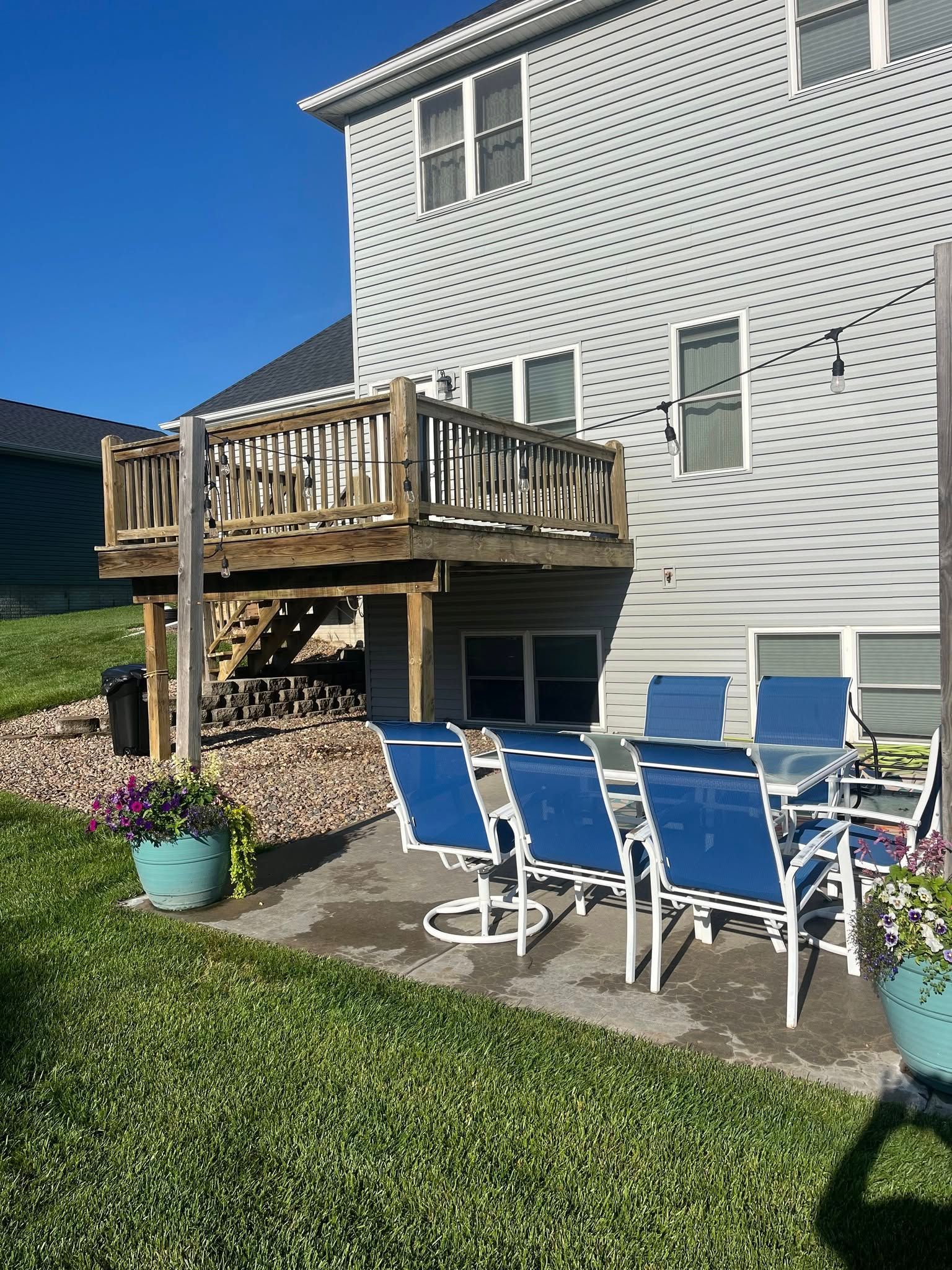 A wooden deck sits above a backyard patio with a blue table set and chairs, set against the side of a gray house.