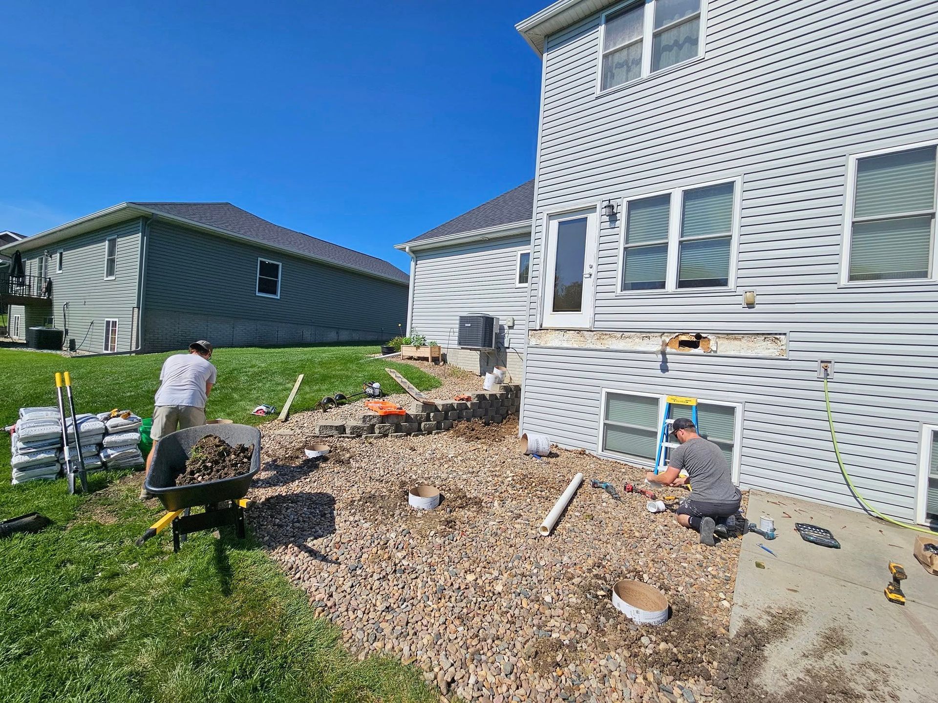 Two workers perform home exterior renovations near a house with gray vinyl siding on a sunny day.