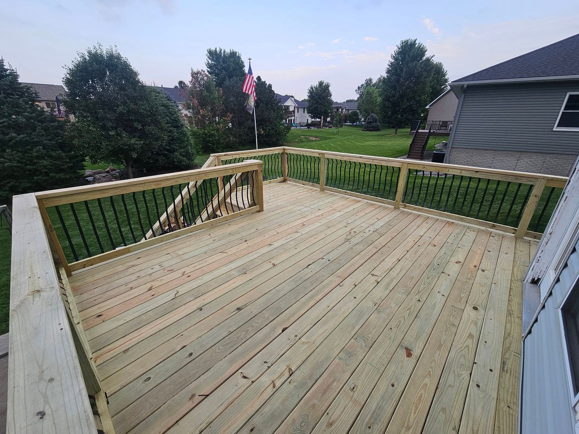 A new wooden deck with railings, overlooking a grassy yard with trees and a house on a sunny day.