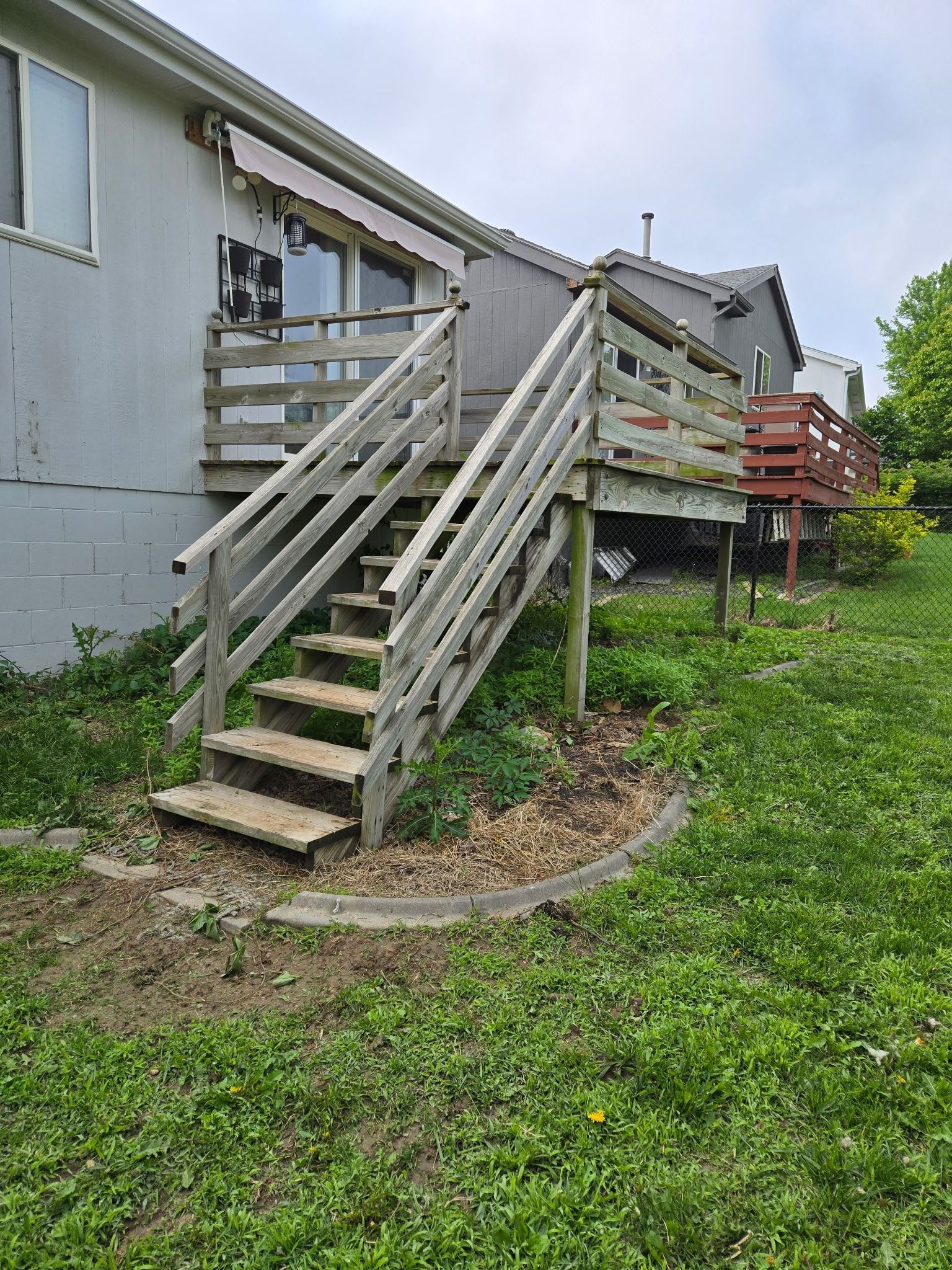 A weathered wooden deck and staircase attached to the back of a light-colored house, situated above a grassy yard.