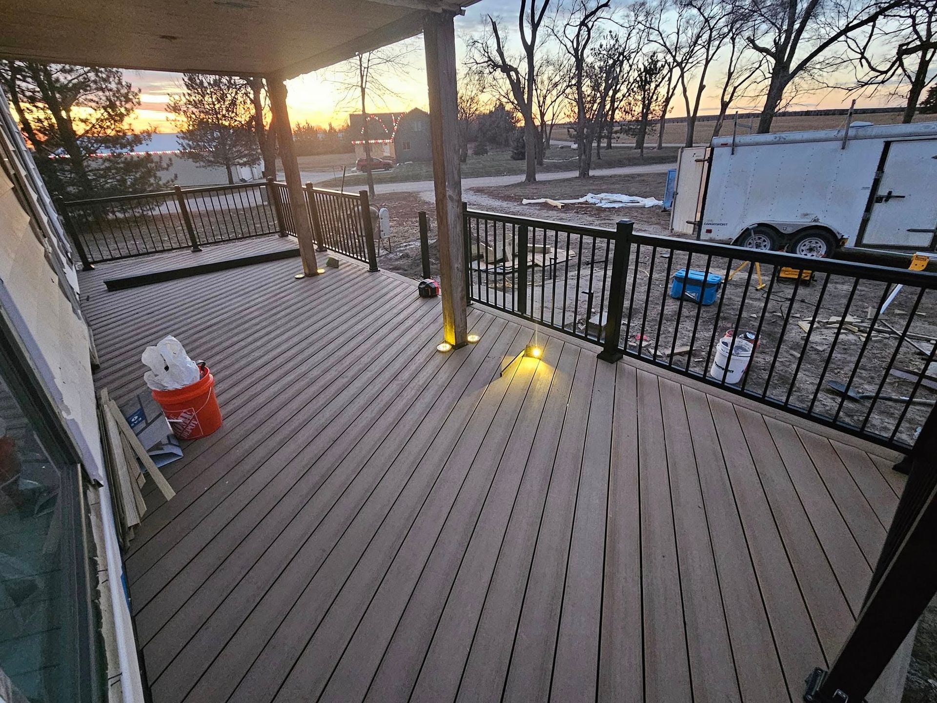 A wooden deck with a black metal railing and built-in lighting during sunset, overlooking a yard with a parked trailer.
