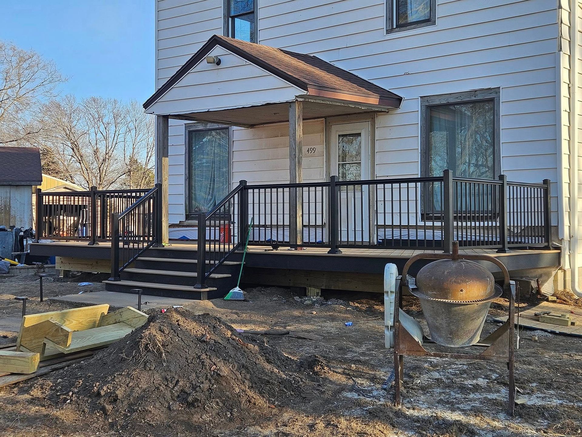 A house under renovation with a newly built dark-toned deck, black railing, stairs, and a concrete mixer in the foreground.