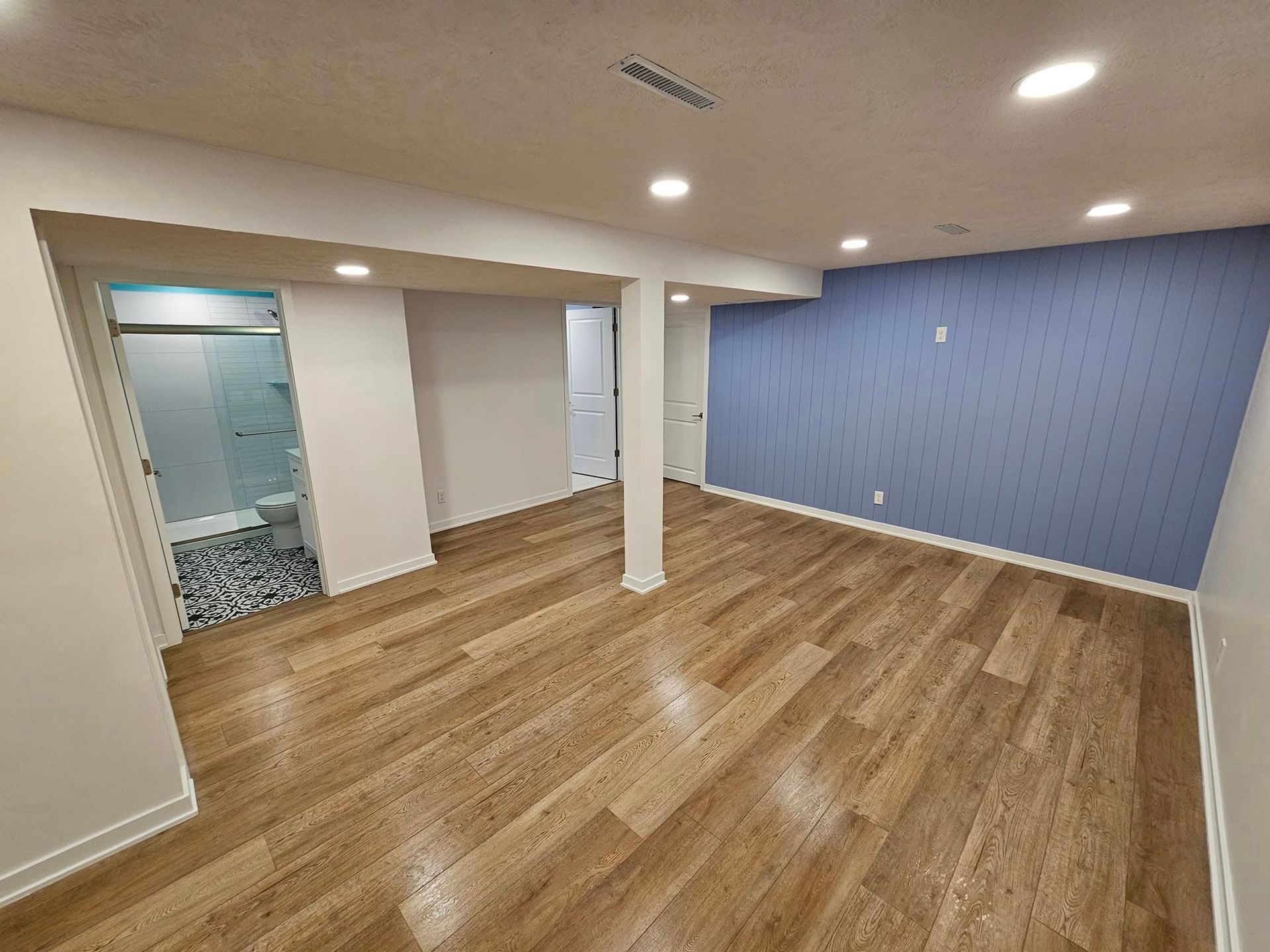 A modern, unfurnished basement room with wood-look flooring, recessed lighting, and one blue accent wall.