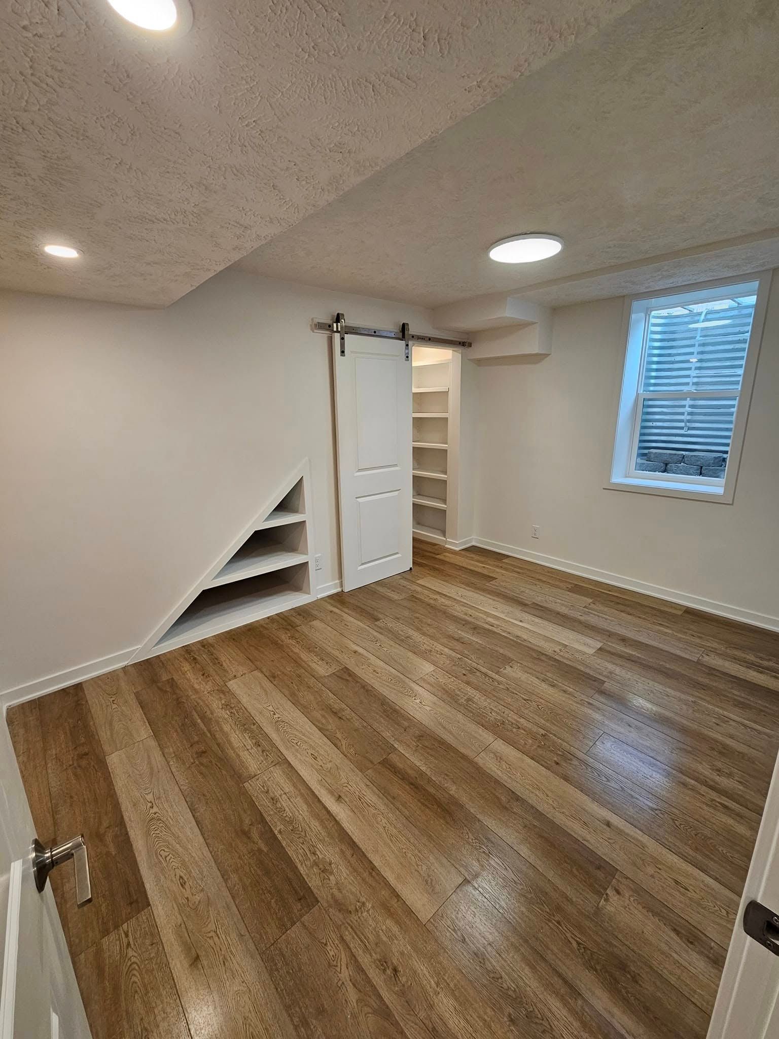 An empty room with light wood floors, a slanted built-in shelf, a white barn door, and a single window with blinds.