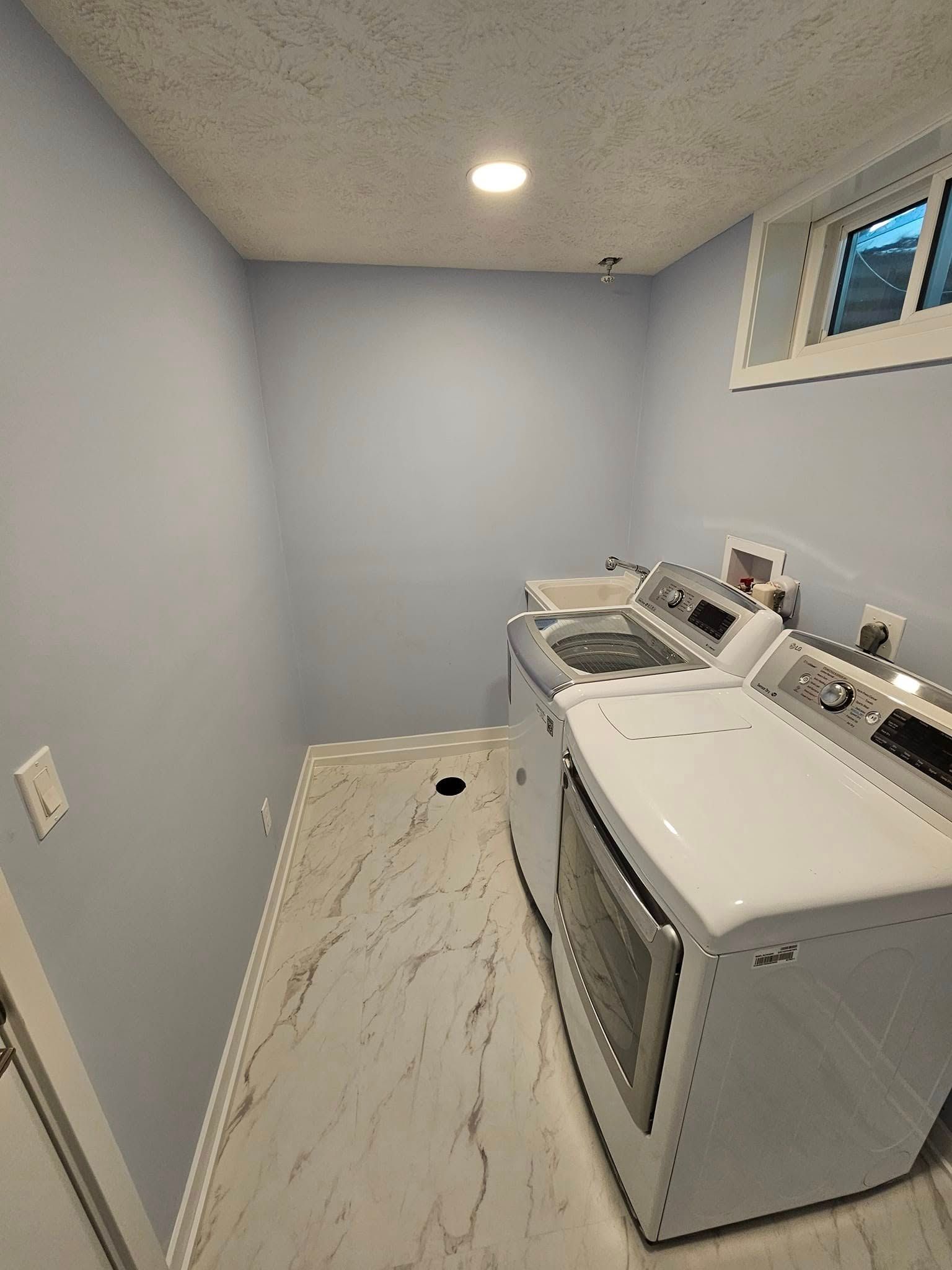 A laundry room with light blue walls, white marbled vinyl flooring, and a white washer and dryer set under a small window.