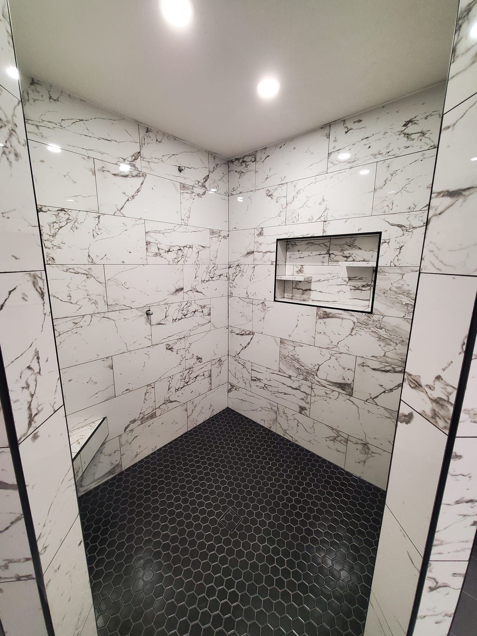 A walk-in shower with white marble-patterned wall tiles, a built-in recessed shelf, and small dark hexagonal floor tiles.