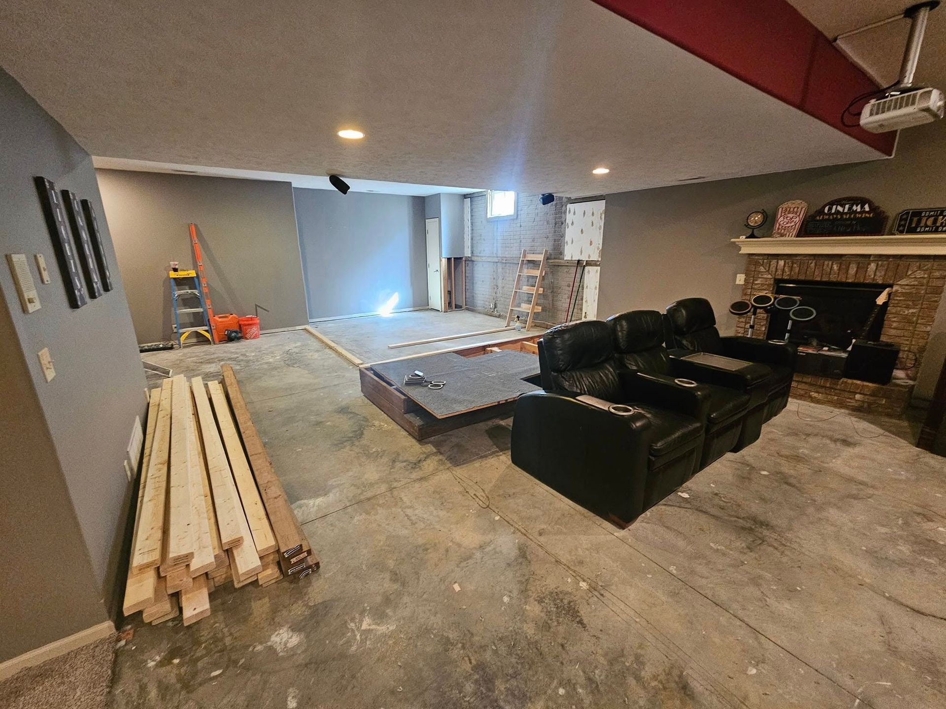 A basement renovation in progress, featuring a pile of lumber, black reclining furniture, and a brick fireplace.