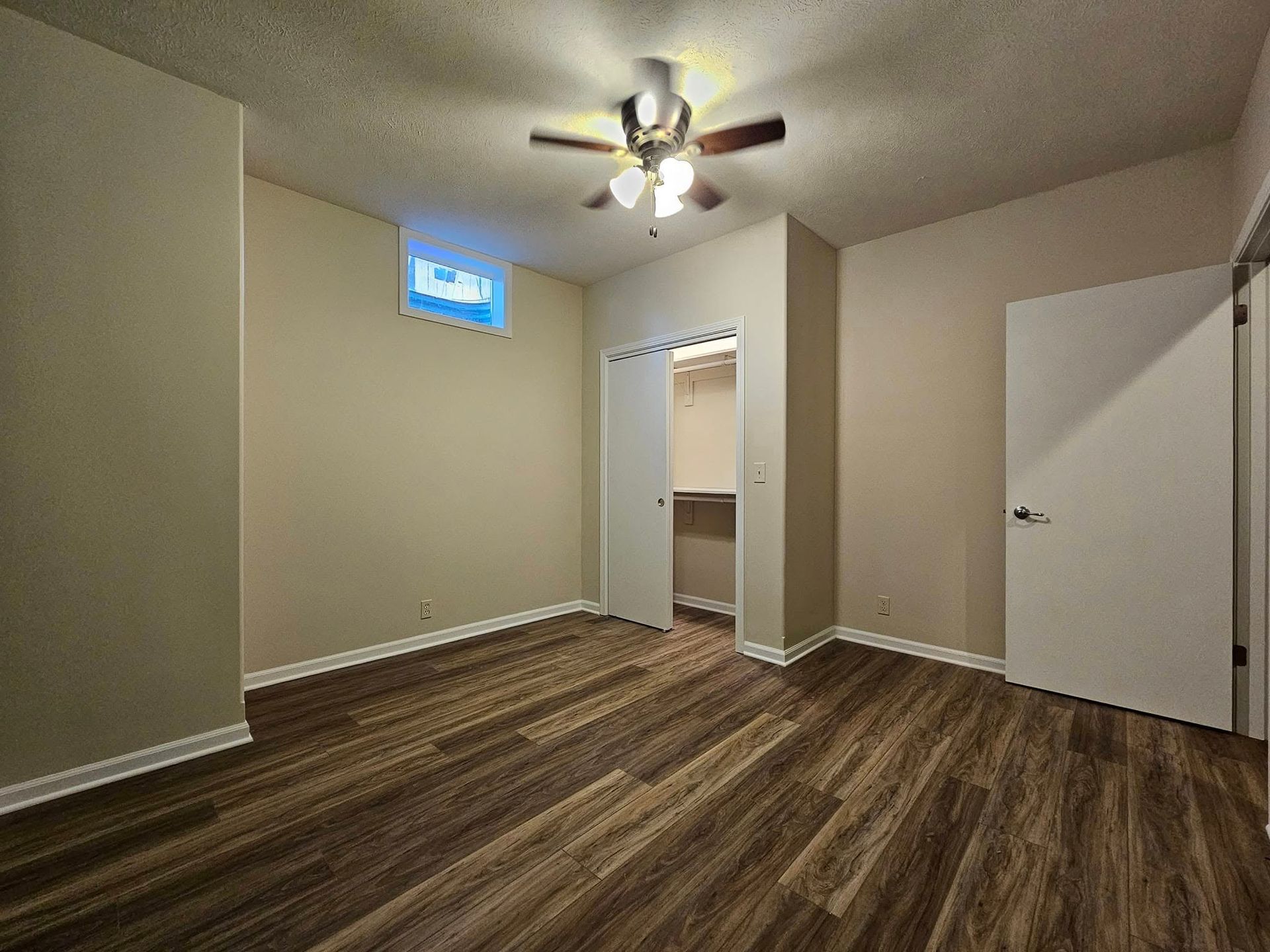 An empty room with light tan walls, a wood-look floor, a ceiling fan, a small high window, and two closed white doors.