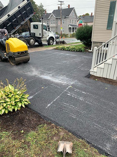 A man is riding a roller on a driveway next to a dump truck.