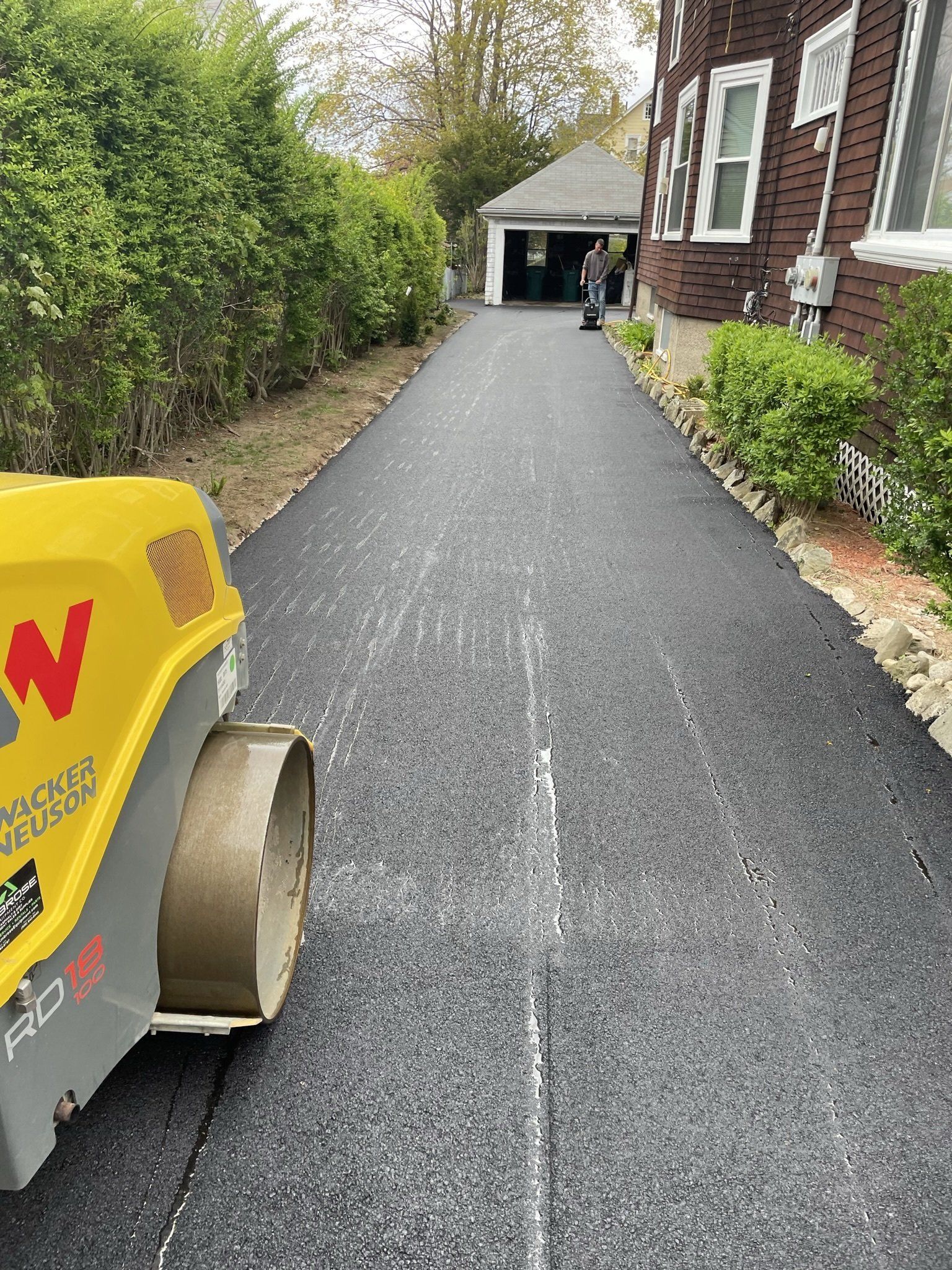 A yellow roller is rolling asphalt on a driveway in front of a house.