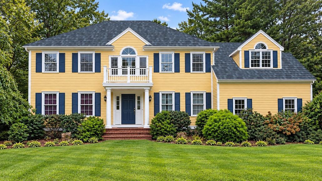 Large yellow colonial-style house with white trim, dark roof, and manicured green lawn under trees