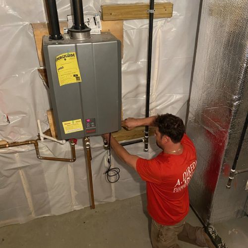 Plumber in red shirt installing a gray tankless water heater on a basement wall with copper pipes