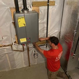 Plumber in red shirt installing a gray tankless water heater on a basement wall with copper pipes