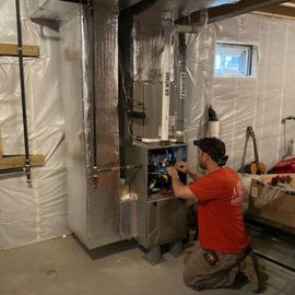 Man in red shirt kneels, working on a furnace in a basement
