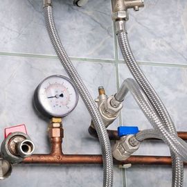 Close-up of plumbing: pressure gauge, pipes, valves, and braided hoses against a tiled wall