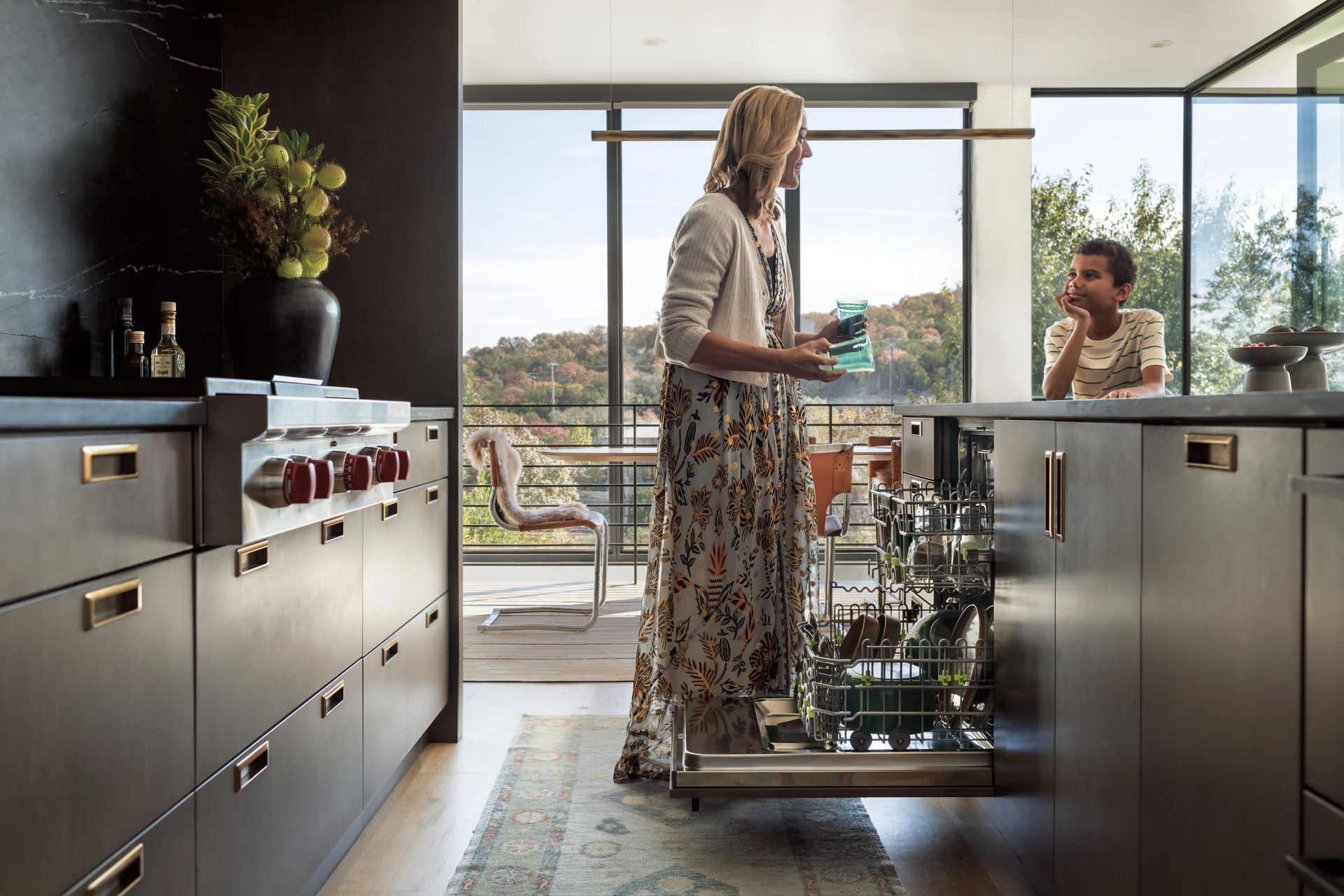 Woman loading a dishwasher in a modern kitchen, child watches, bright landscape visible through the windows.
