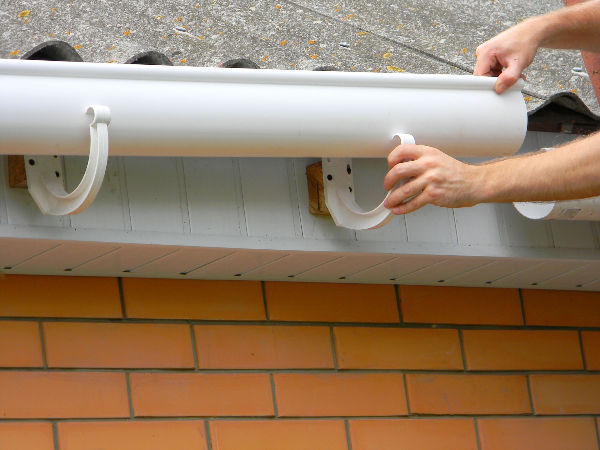 Hands mounting a white gutter onto brackets attached to the fascia board of a brick building.