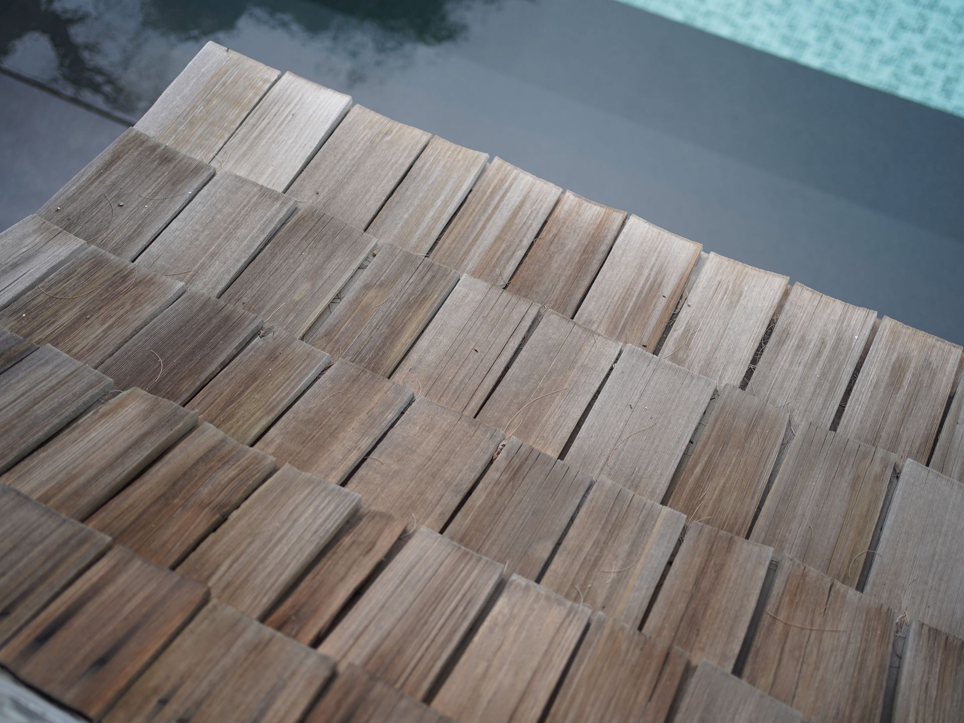 An angled, close-up view of weathered, overlapping cedar wood shingles on a roof edge, with a pool visible in the background.