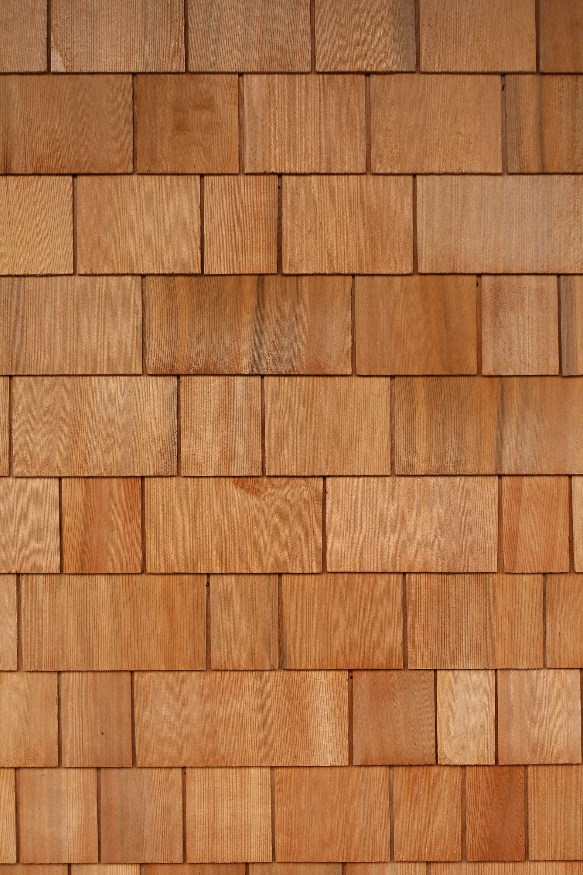 A close-up view of a wall covered in light brown wooden shingles arranged in a repeating, staggered horizontal pattern.