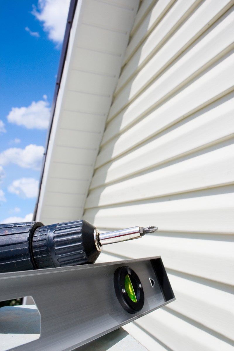 A close-up view of a power drill and a spirit level positioned against beige vinyl siding on a house exterior.
