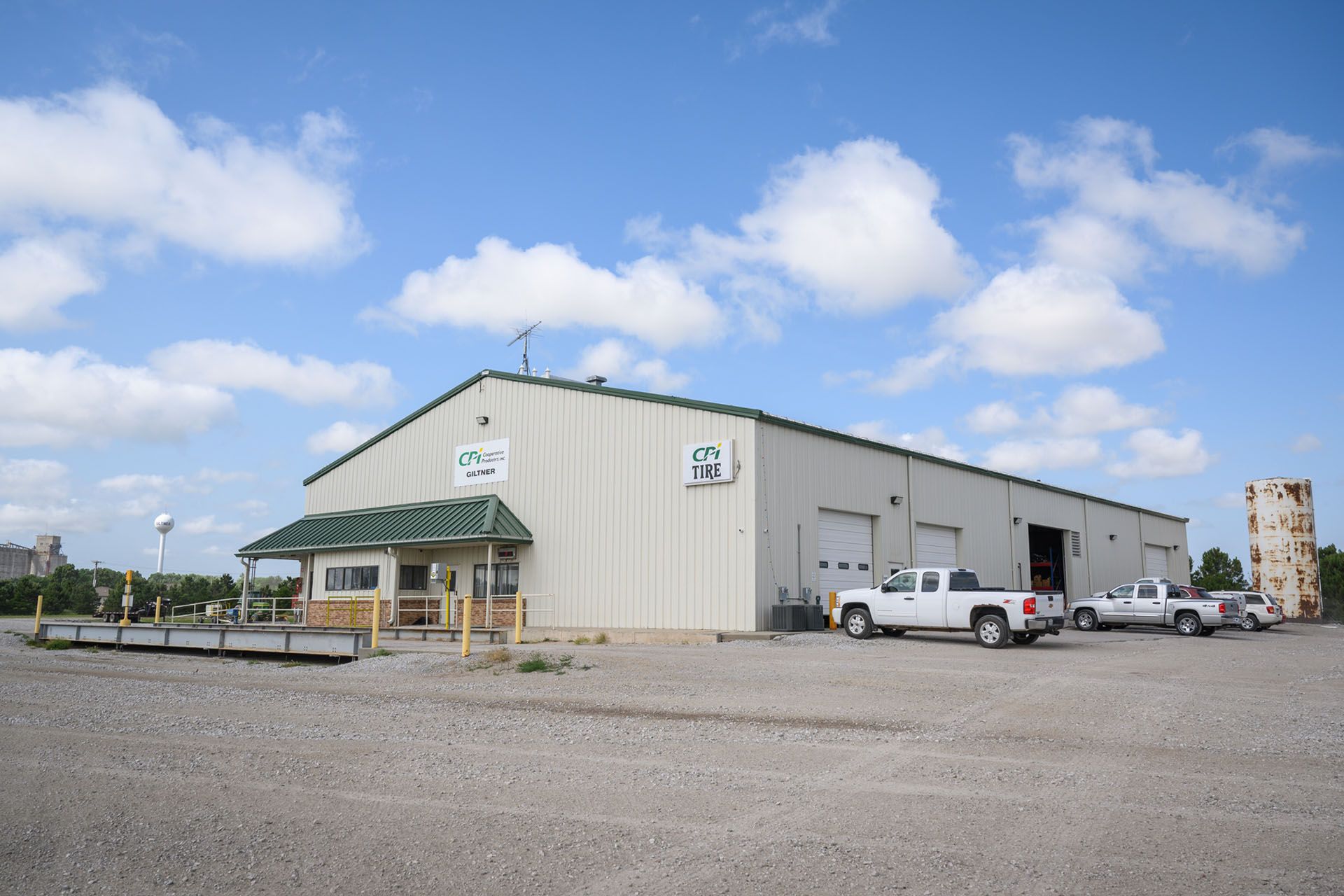 Large beige warehouse with loading dock, parked pickup trucks, and a gravel lot under a blue sky