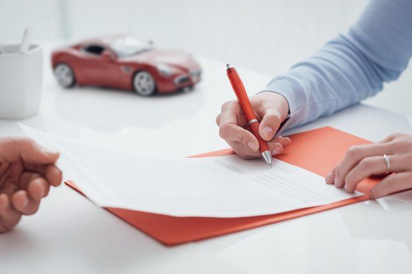 A person signs a document on an orange folder on a desk, with a red toy car and white mug nearby.