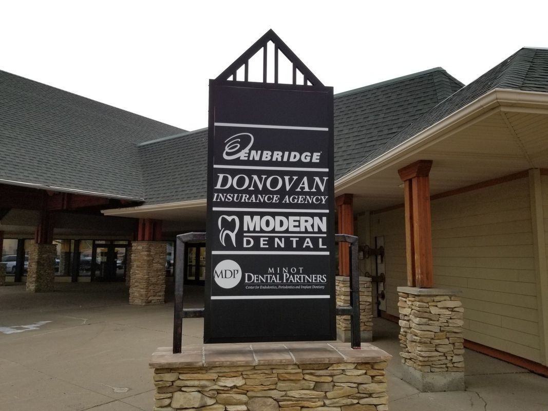 A multi-tenant black sign stands on a stone base in front of a building with a gray roof and tan siding.