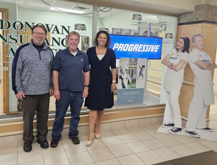 Three people stand smiling in front of a Donovan Insurance Agency office with a Progressive display and cardboard cutouts.