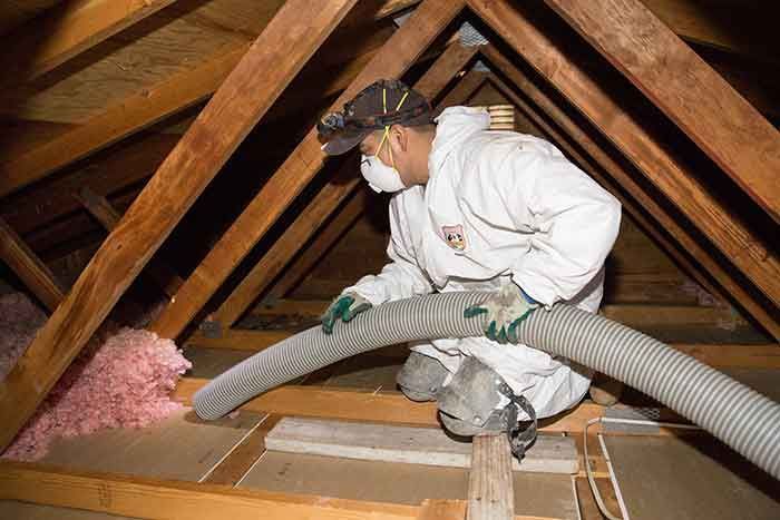 Worker in white protective suit spraying insulation into an attic with exposed wooden beams.