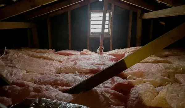 Attic with pink insulation and a small window. Wooden beams frame the space; sunlight streams in.