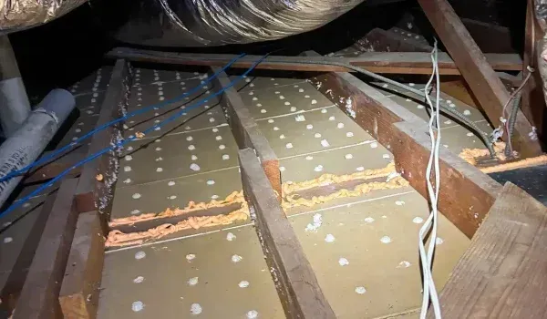 Attic interior showing wood beams, insulation, and wiring. Orange sealant visible between insulation panels.